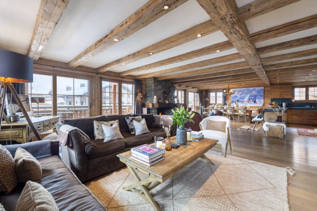 Spacious living room with exposed wooden beams, large windows, a dark sofa, coffee table with books, cozy chairs, dining area, fireplace, and open kitchen in a bright, rustic interior. David Pearson Travel