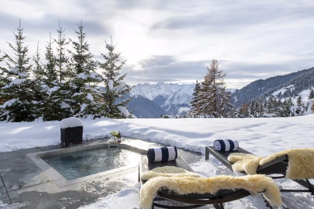 A snowy mountain landscape with a small outdoor hot tub, two lounge chairs with fur throws and rolled towels, surrounded by pine trees under a cloudy sky. David Pearson Travel