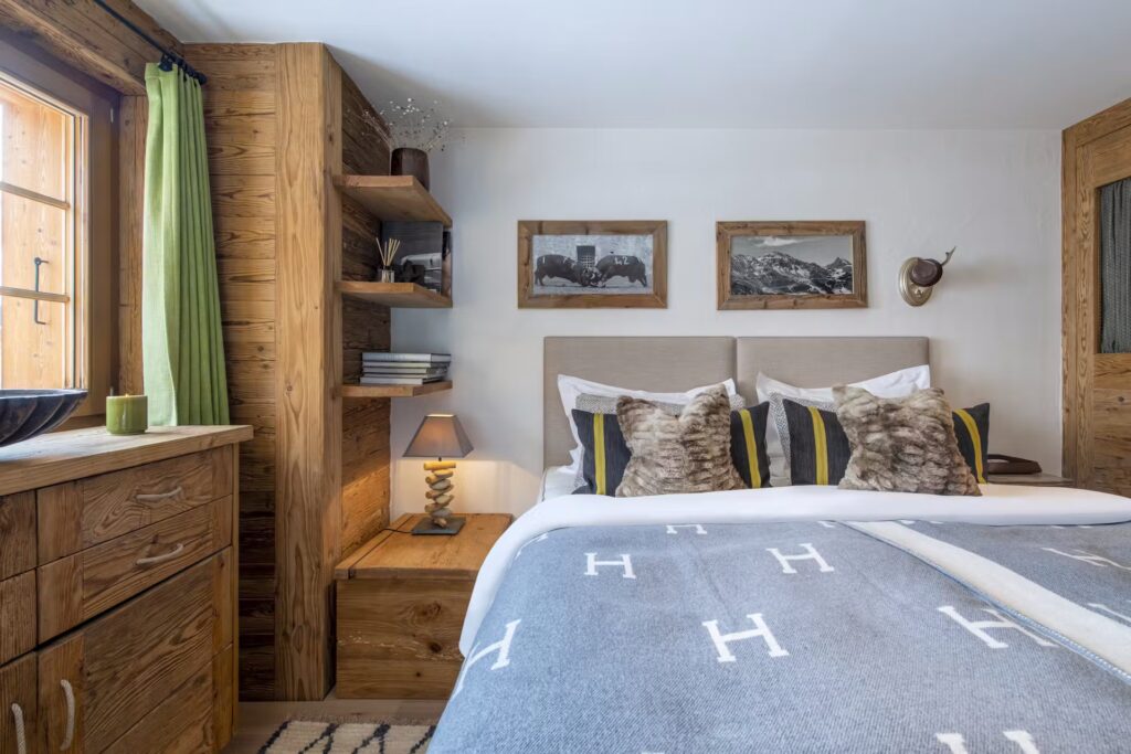 A cozy bedroom with wood-paneled walls and furniture, a bed with decorative pillows and a grey blanket, two framed photos on the wall, a small lamp, open shelves with books, and a window with green curtains. David Pearson Travel