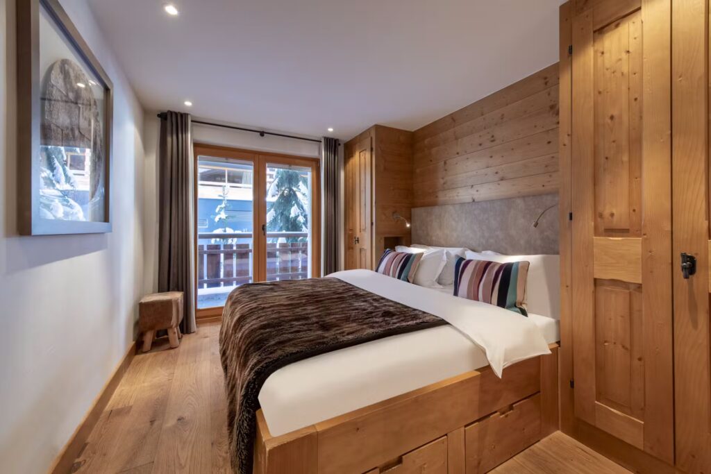 Cozy bedroom with wooden walls and floors, a large bed with brown and white bedding, striped pillows, soft lighting, a framed picture on the wall, and glass doors leading to a snowy balcony. David Pearson Travel