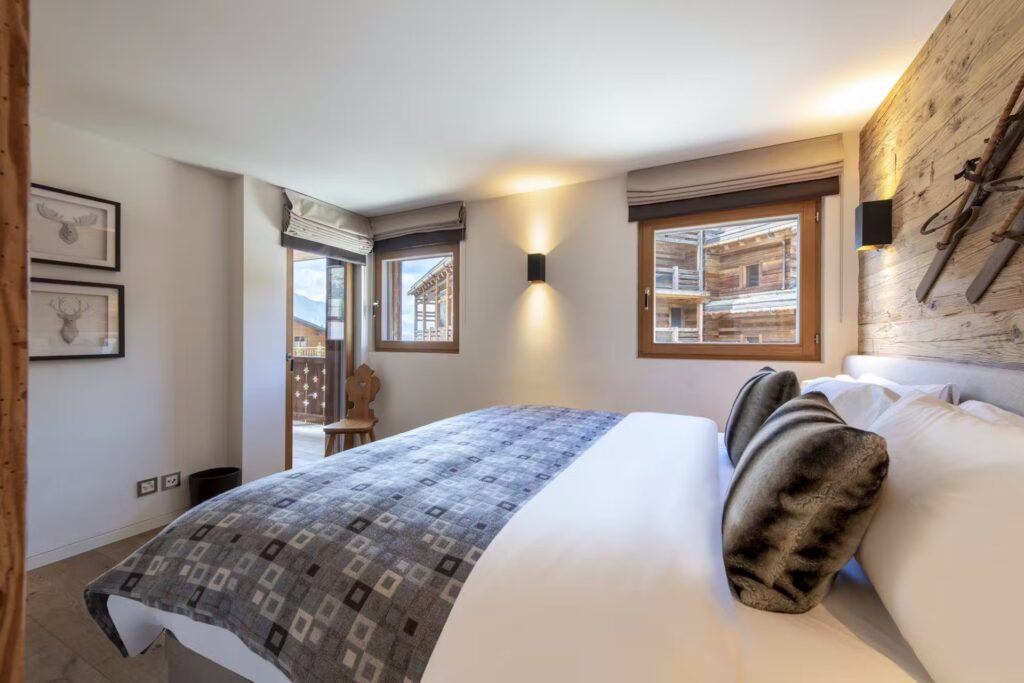 A cozy bedroom with a large bed, patterned throw, and two dark pillows, featuring wooden accents, framed art, a faux fur chair, and windows overlooking chalets outside. David Pearson Travel