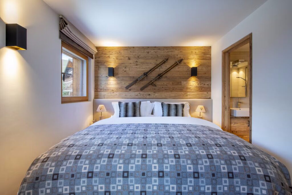 A cozy bedroom with a large bed, geometric-patterned blue-grey bedding, wood-paneled accent wall with vintage skis, bedside lamps, a window with a shade, and an open bathroom door to the right. David Pearson Travel