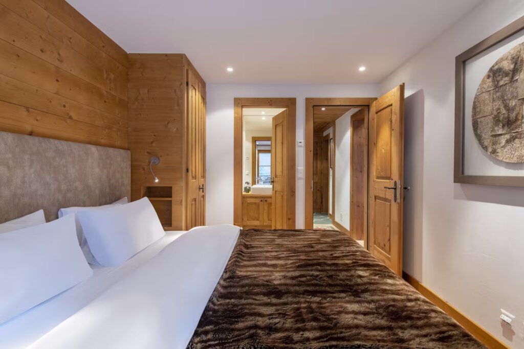 A cozy bedroom with a large bed topped with white linens and a brown fur blanket, wooden walls and doors, built-in wardrobe, soft lighting, and a framed round artwork on the white wall. David Pearson Travel