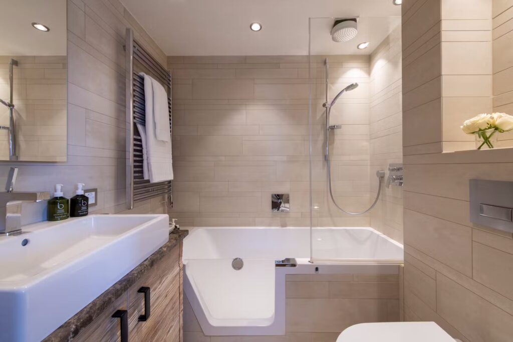 A modern bathroom with beige tiles, a white sink on a wooden cabinet, a bathtub with a glass shower screen, wall-mounted showerhead, towels on a rack, and a vase of white flowers on a ledge. David Pearson Travel
