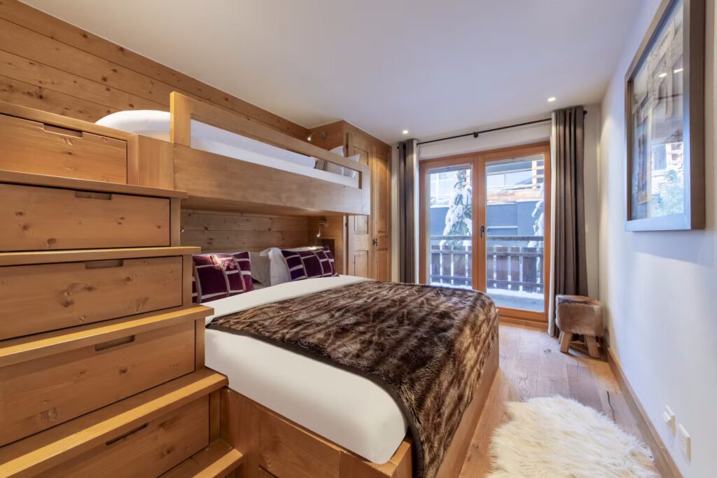 A cozy bedroom with wooden bunk beds, a large bed below with a fur blanket, wooden stairs, soft lighting, a small stool, a fluffy white rug, and a glass door leading to a balcony with mountain views. David Pearson Travel