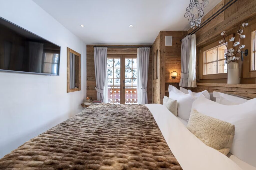 A cozy bedroom with a large bed covered in a brown fur blanket, white pillows, wood-paneled walls, a wall-mounted TV, and glass doors opening to a balcony with snowy trees outside. David Pearson Travel