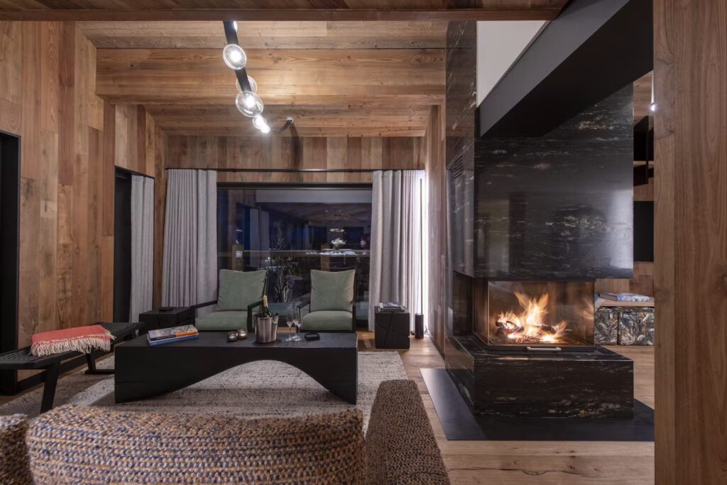 A modern, cozy living room with wood-paneled walls, a black stone fireplace with a fire burning, green armchairs, a dark coffee table, and a large window with curtains showing a night view outside. David Pearson Travel
