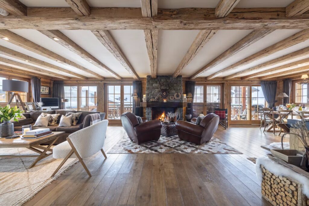 A spacious, rustic living room with exposed wooden beams, a stone fireplace, two leather chairs, cozy seating, large windows, and wooden floors, filled with natural light and mountain or forest views outside. David Pearson Travel