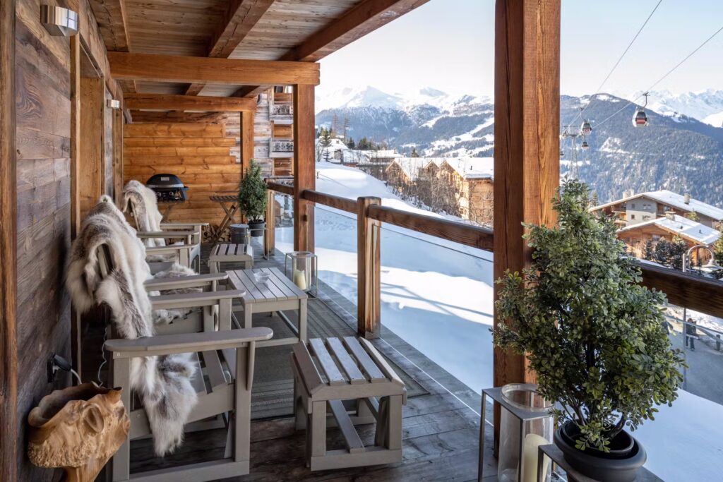 A wooden chalet balcony with fur-covered chairs and potted plants overlooks snowy mountains and ski lifts, with clear skies and distant alpine cottages visible. David Pearson Travel
