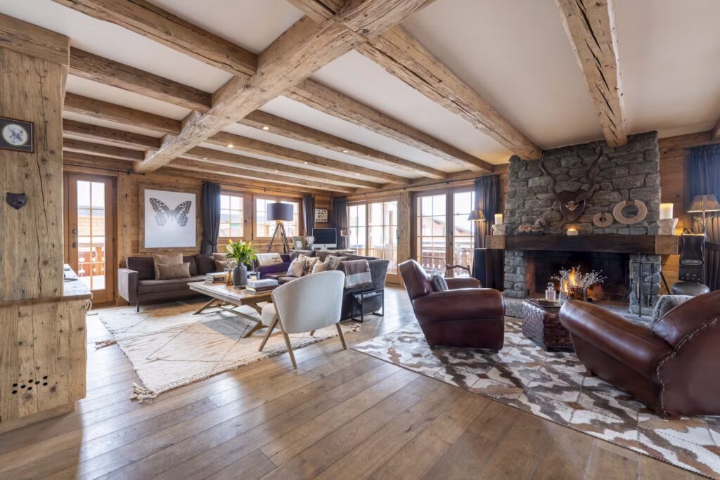 Spacious rustic living room with exposed wooden beams, stone fireplace, leather chairs, modern sofas, large windows, hardwood floors, and cozy décor, creating a warm and inviting atmosphere filled with natural light. David Pearson Travel