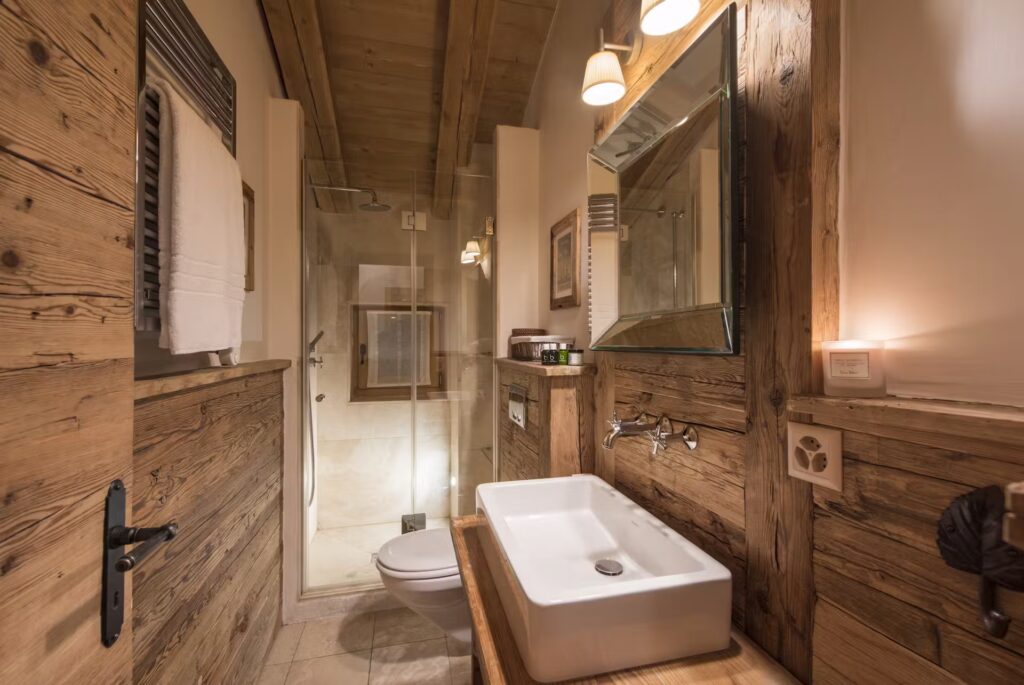 A cozy rustic bathroom with wooden walls, a white rectangular sink, a large mirror, modern fixtures, a glass shower enclosure, and warm lighting creating a welcoming atmosphere. David Pearson Travel