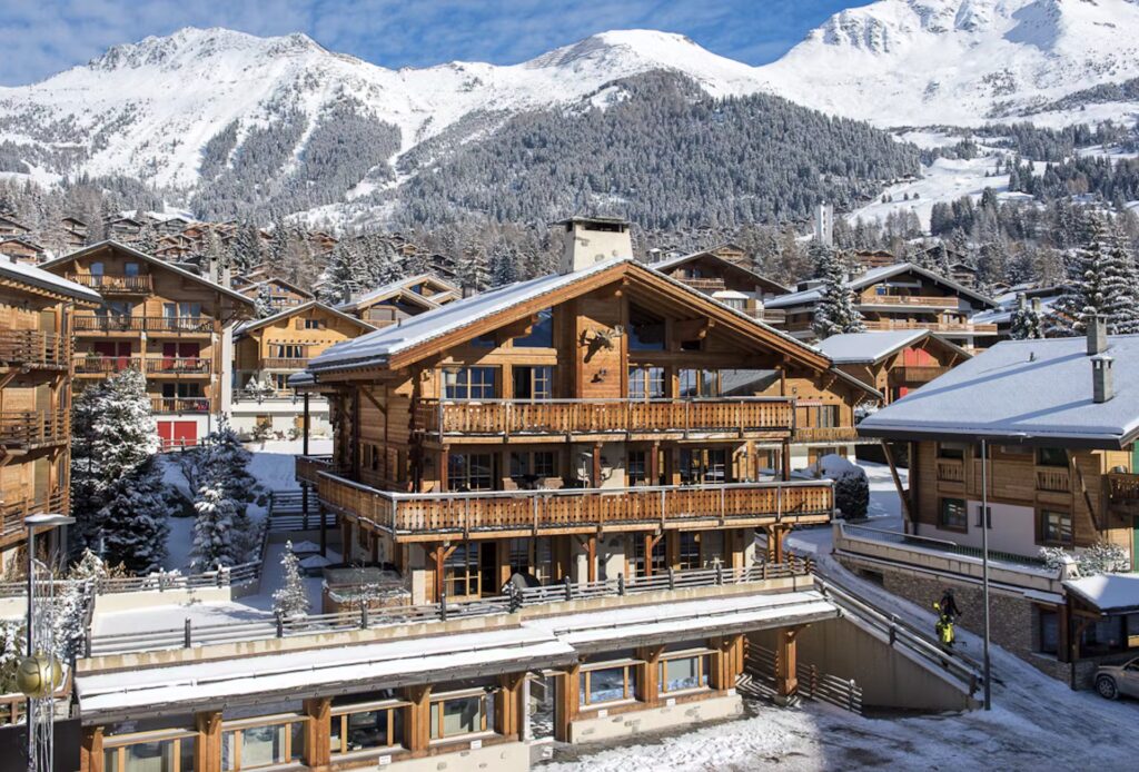 A large wooden chalet with balconies stands among snowy rooftops in a mountain village, surrounded by snow-covered trees and tall, white-peaked mountains under a blue sky. David Pearson Travel