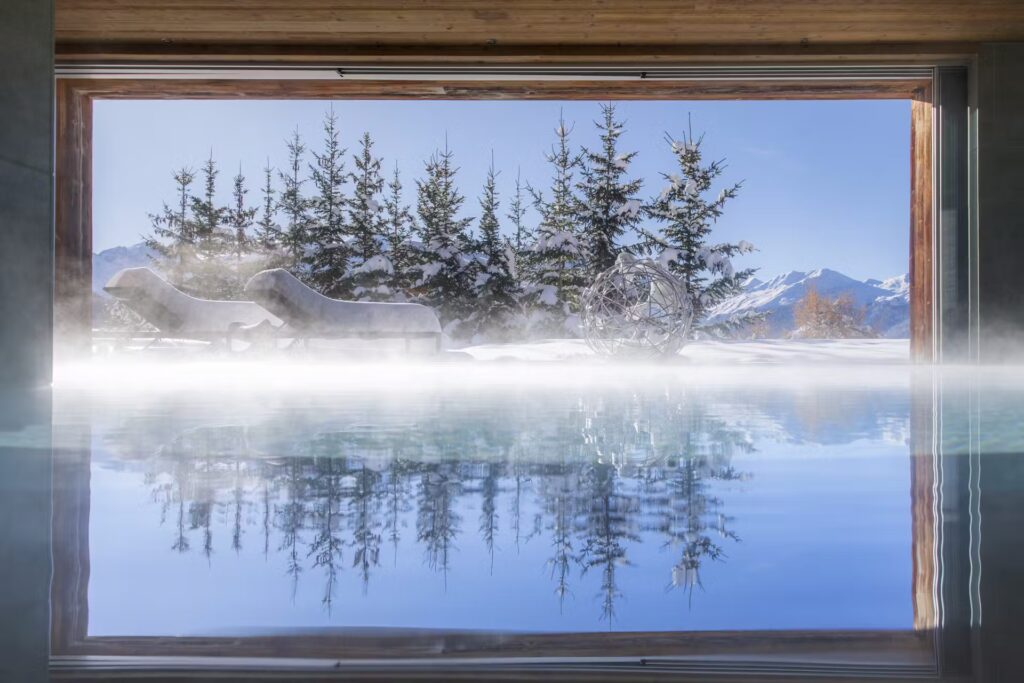 An indoor pool opens to a snowy outdoor scene with frosted lounge chairs, pine trees, and mountains in the background. Steam rises from the water, blending with the winter landscape. David Pearson Travel