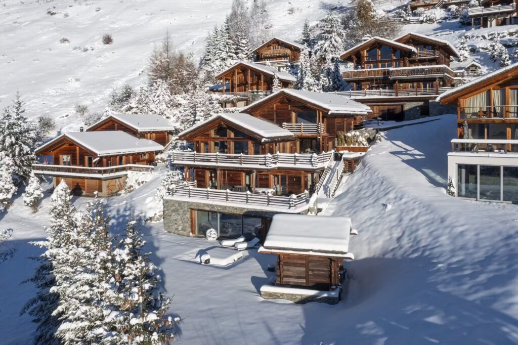 A cluster of wooden chalets with large balconies sits on a snowy hillside, surrounded by snow-covered trees and fresh snow, under a clear blue sky. David Pearson Travel