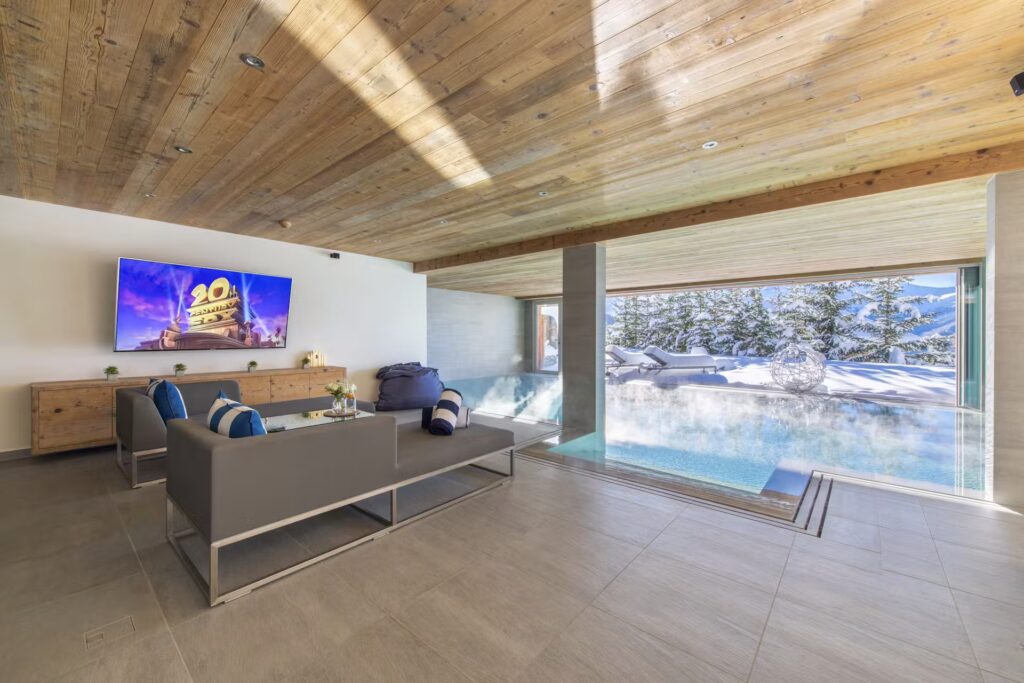 A modern living room with gray sofas, a flat-screen TV, and a wooden ceiling, next to an indoor pool with large windows overlooking a snowy, pine-filled landscape. David Pearson Travel