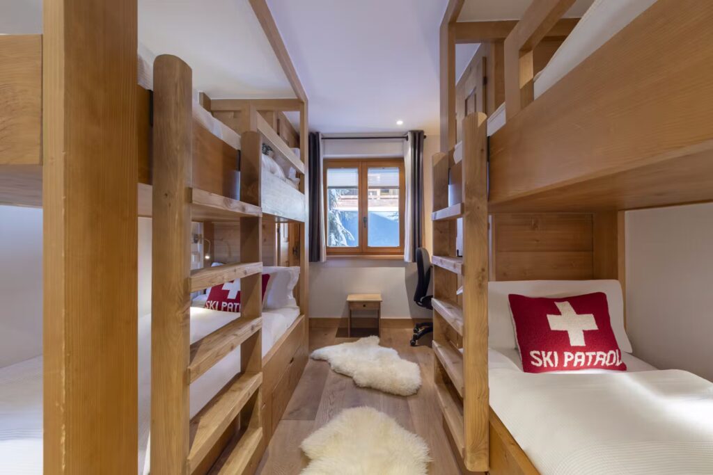 A cozy room with four wooden bunk beds, each with white bedding and red Ski Patrol pillows. Two white furry rugs are on the wooden floor, and a window with mountain views lets in natural light. David Pearson Travel