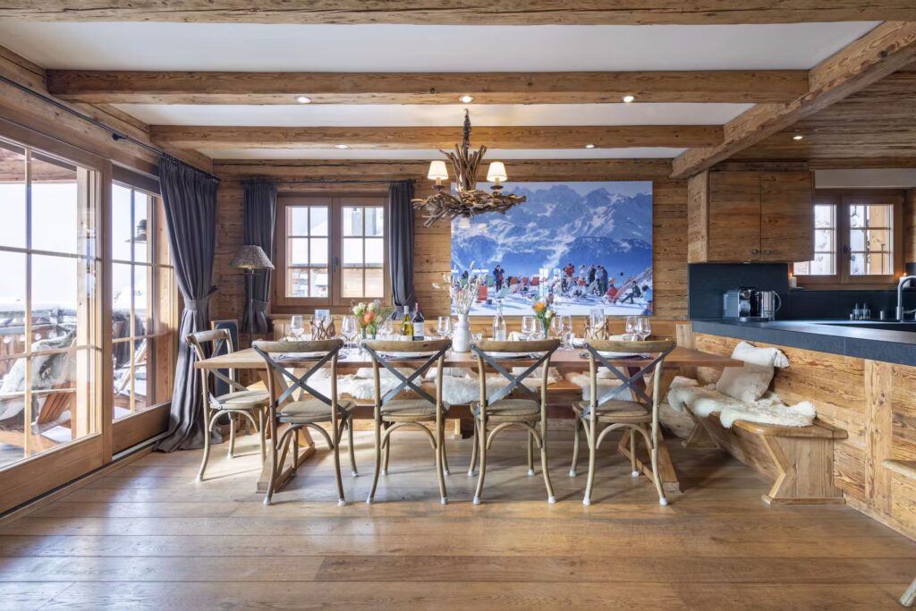 A rustic dining room with a long wooden table, ten chairs, sheepskin bench, and a mountain scene photo on the wall. Large windows let in natural light; wood beams and floors create a cozy chalet ambiance. David Pearson Travel