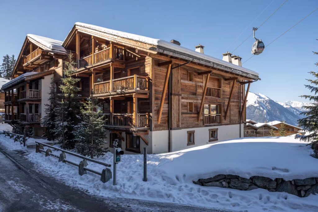 A wooden chalet-style building stands amid snow with balconies overlooking a wintry landscape. Pine trees and a snow-covered road are in front, and a cable car moves overhead against a backdrop of mountains under a clear blue sky. David Pearson Travel