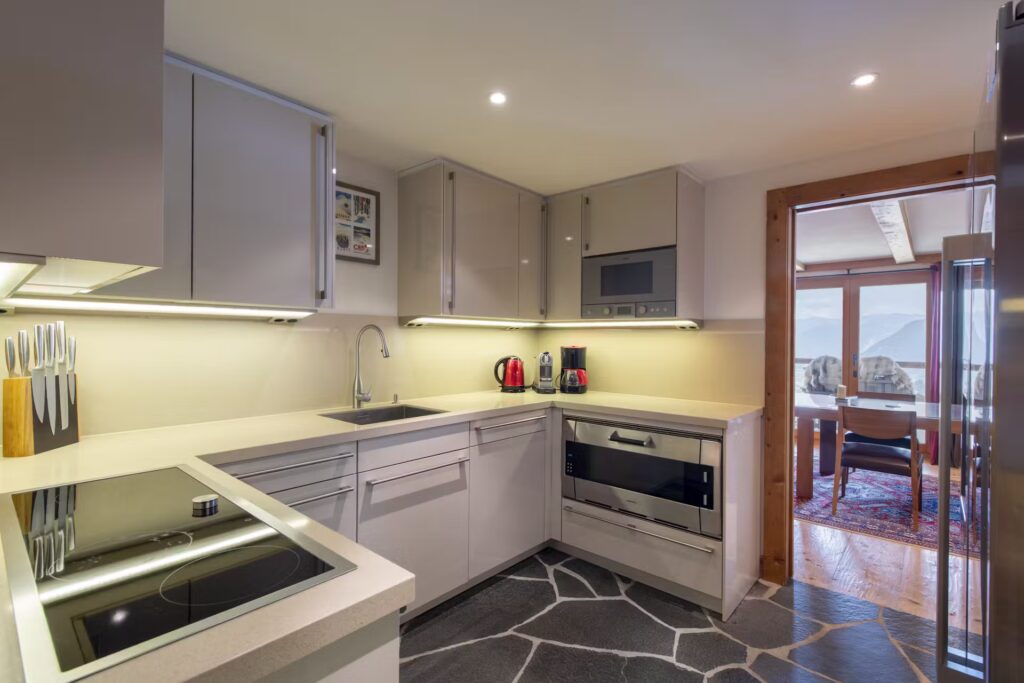 Modern kitchen with light-colored cabinets, built-in appliances, under-cabinet lighting, a set of knives, and a sink. The kitchen opens into a dining area with large windows and scenic views. David Pearson Travel