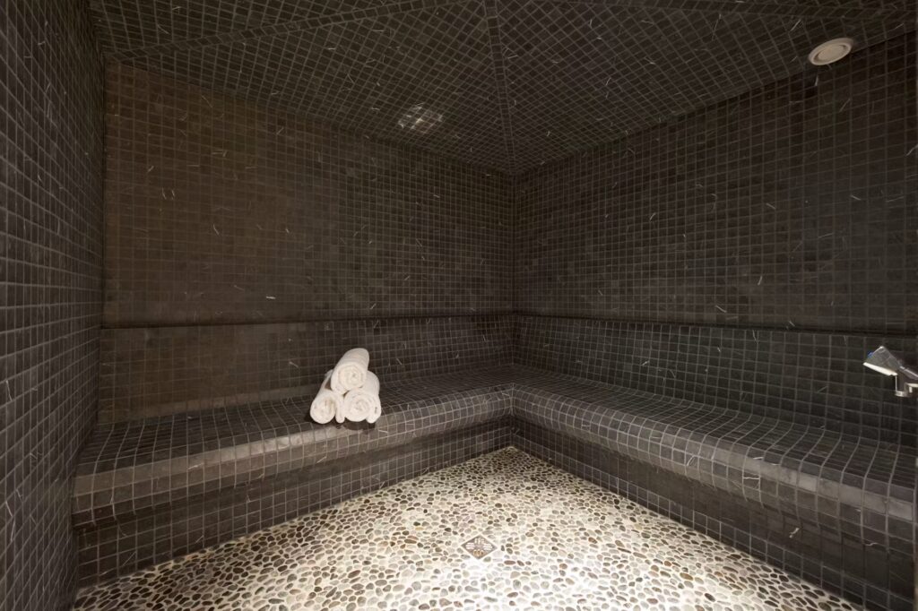A modern steam room with dark tiled walls and benches, a pebble-patterned floor, and a stack of rolled white towels placed on one of the benches. David Pearson Travel