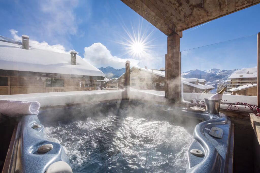 A steaming hot tub on a snowy balcony overlooks alpine rooftops and distant mountains, with bright sunshine and a blue sky above. A towel and champagne bucket are set on the tub’s edge. David Pearson Travel