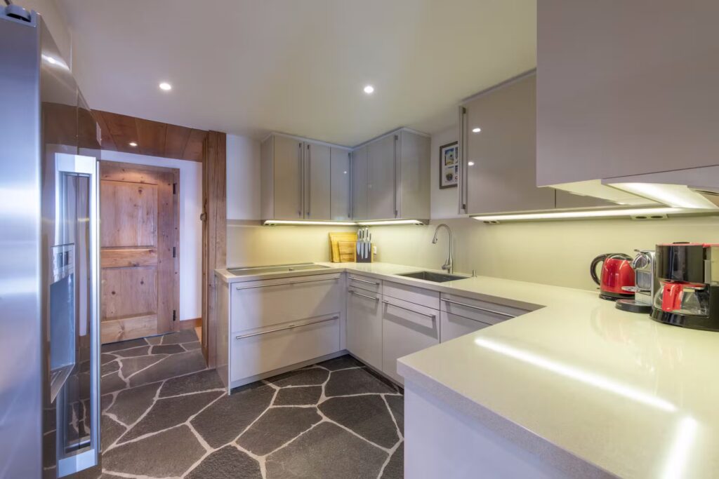 Modern kitchen with light-colored cabinets, stone-patterned floor, built-in appliances, countertop with a coffee maker, toaster, and kettle, under-cabinet lighting, and a wooden door in the background. David Pearson Travel