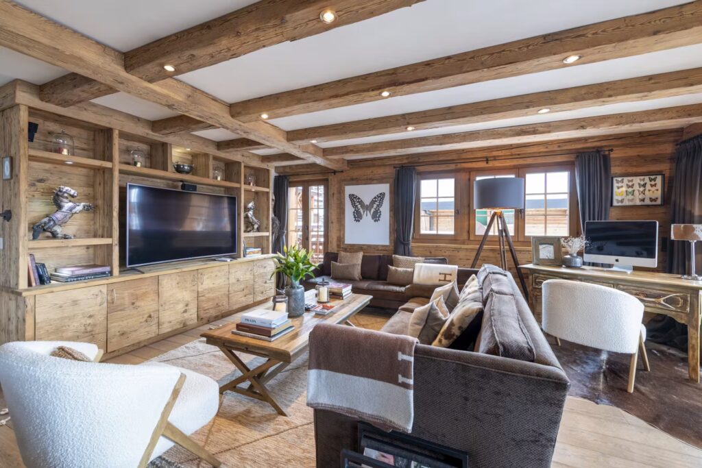 Cozy living room with wooden beams, built-in shelves, modern art, a large TV, brown sofas, white armchairs, a coffee table, desk with a computer, and large windows letting in natural light. David Pearson Travel
