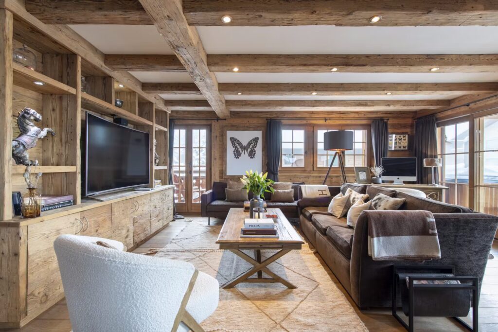 A cozy living room with rustic wooden beams, large windows, a mounted TV, shelves, a butterfly artwork, modern and classic furniture, and a coffee table with books and flowers. Natural light fills the space. David Pearson Travel