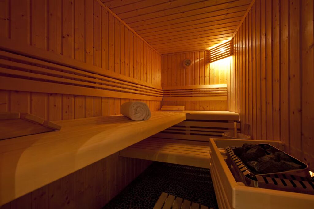 A wooden sauna room with warm lighting, two-tiered benches, a rolled-up towel on the upper bench, and a heater with sauna stones on the right side. David Pearson Travel
