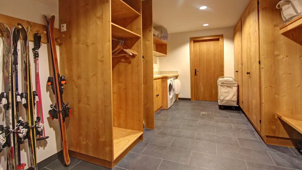 A mudroom with wooden lockers, hooks, and open shelves, ski and snowboard gear stored on the left, a washing machine, beige suitcase, and tiled dark gray floor. David Pearson Travel