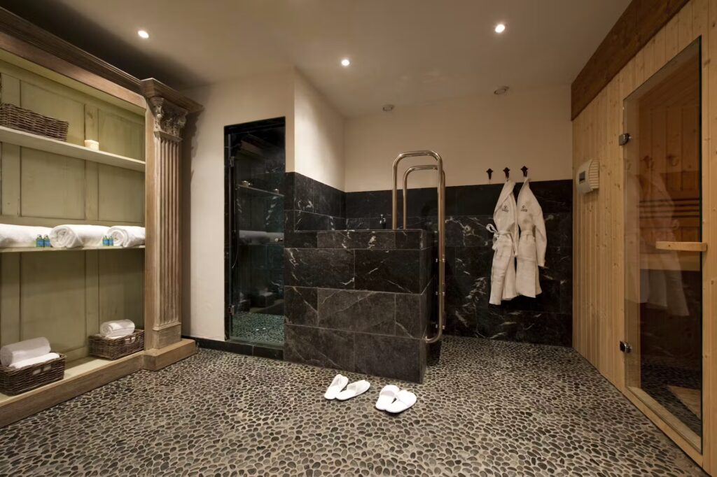 A luxurious spa room with a pebble stone floor, open shelving with towels, a black marble shower area, wooden sauna, two white robes on hooks, and white slippers on the floor. David Pearson Travel