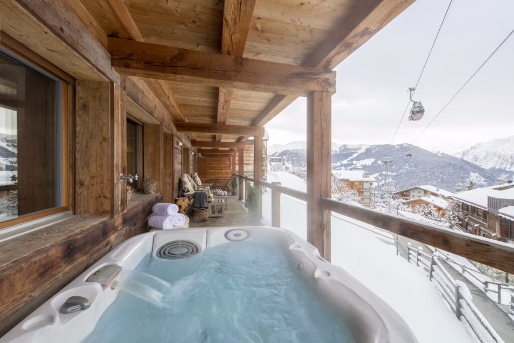 A hot tub on a wooden balcony overlooking snowy mountains and a ski lift, with folded towels and lounge chairs nearby, creating a cozy and relaxing winter scene. David Pearson Travel