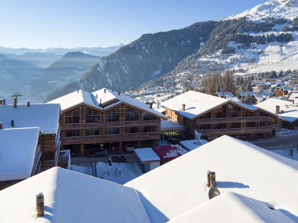Snow-covered chalet rooftops overlook a scenic mountain valley on a clear, sunny winter day. Wooden buildings and evergreen trees dot the snowy landscape, with mountain slopes in the background. David Pearson Travel