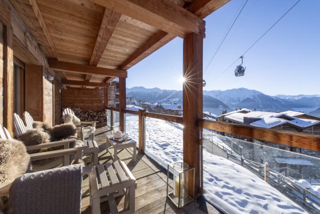 A wooden chalet balcony with cozy chairs overlooks snowy mountains and rooftops. A cable car passes by in the blue sky, and sunlight shines through clear glass railings. Firewood is stacked nearby. David Pearson Travel