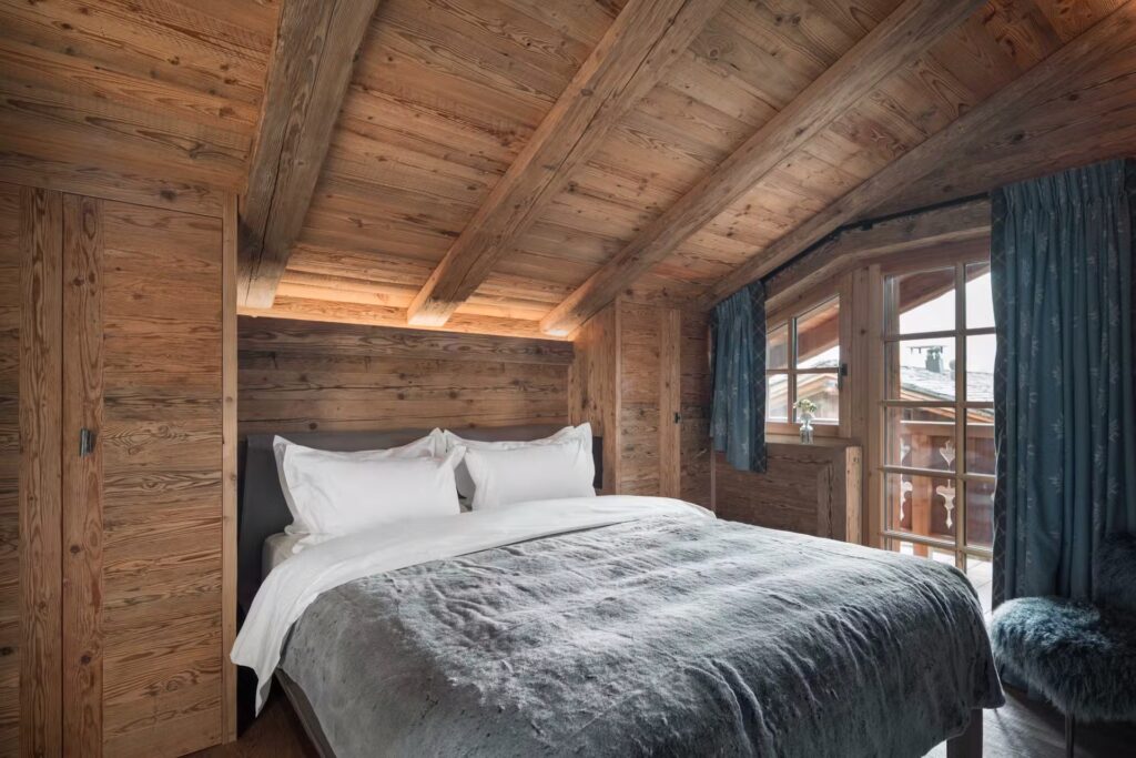A cozy bedroom with rustic wooden walls and ceiling, a neatly made bed with white linens and a gray blanket, blue curtains, and a window letting in natural light, offering a view of a wooden balcony. David Pearson Travel