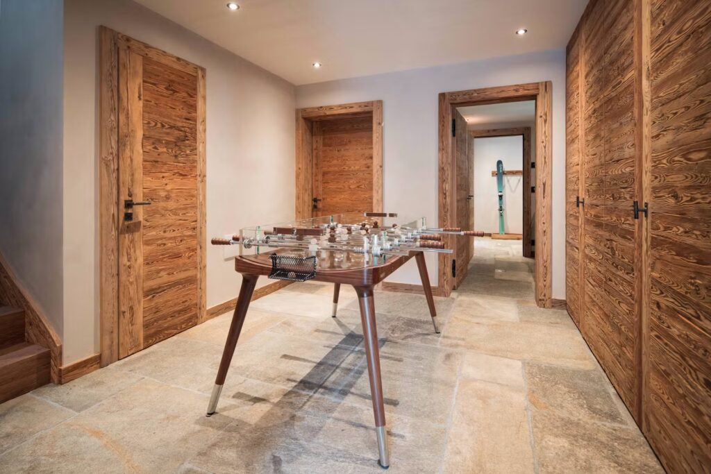A hallway with light stone flooring, wooden doors and walls, and a foosball table in the center; an ironing board is visible through a doorway in the background. David Pearson Travel
