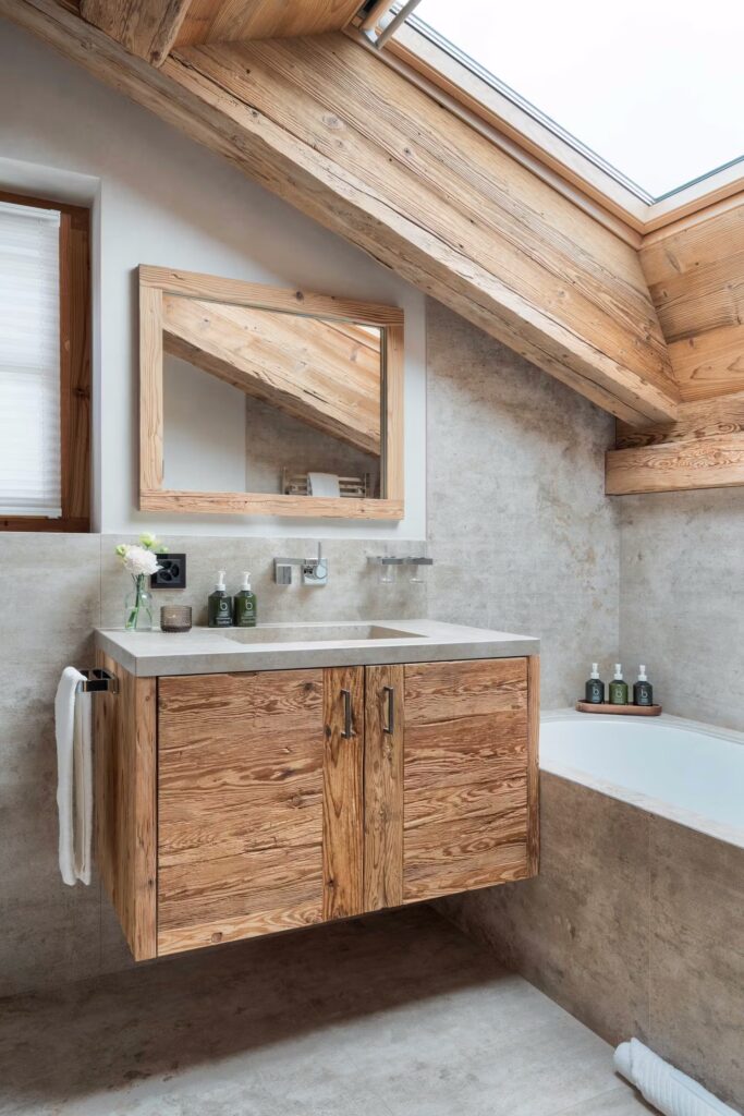 Modern bathroom with a wooden floating vanity, matching mirror, stone countertop, and an integrated bathtub under a sloped wooden ceiling with a skylight. Towels and toiletries are neatly arranged. David Pearson Travel