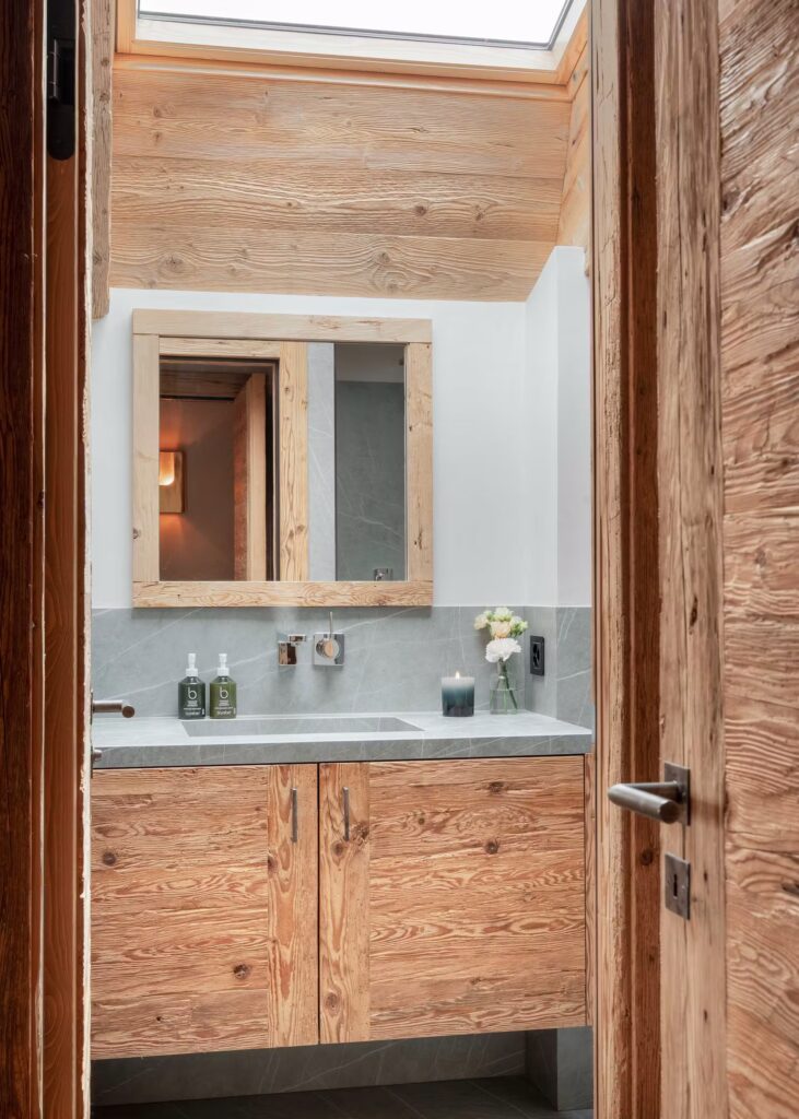 A modern bathroom with natural wood cabinets and door, a gray countertop, a large mirror, a skylight, and minimalist decor including soap dispensers, a candle, and a small vase of flowers. David Pearson Travel