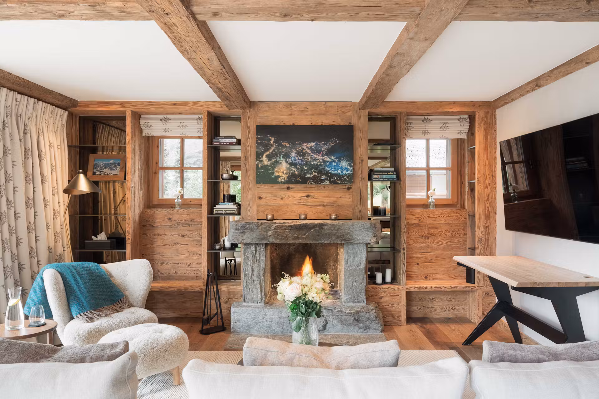 A cozy living room with wooden beams, a stone fireplace with a fire burning, shelves with books and decor, a large wall-mounted TV, a bouquet of flowers on the coffee table, and comfortable seating. David Pearson Travel