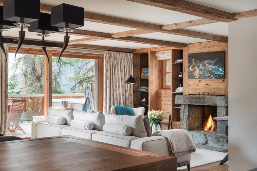 Cozy living room with exposed wooden beams, a stone fireplace, bookshelves, and a white sofa. Large glass doors open to a balcony with outdoor seating and a view of pine trees. Warm, inviting atmosphere. David Pearson Travel