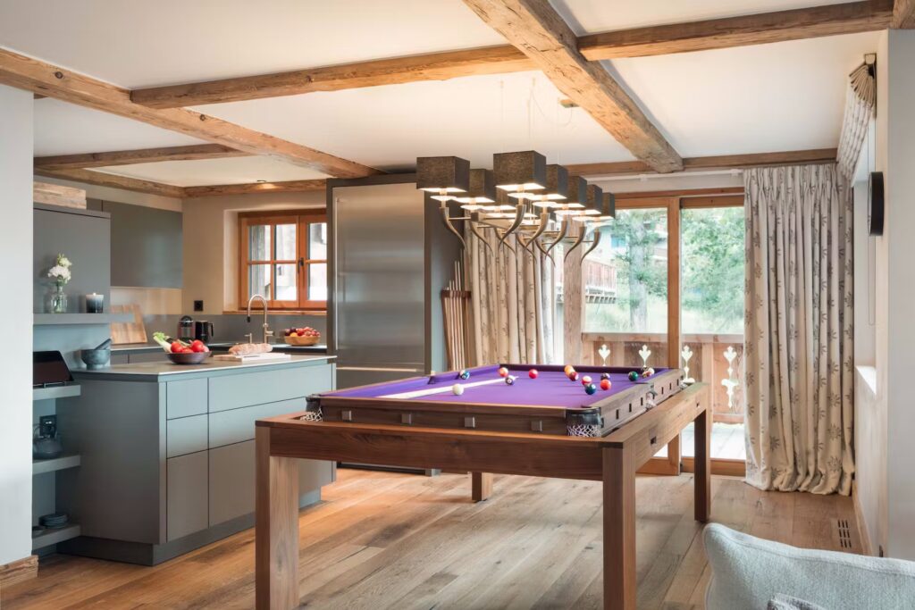 A modern kitchen and dining area with wooden beams, featuring a pool table with a purple felt top in the center, light wood flooring, and large windows with a view of greenery outside. David Pearson Travel