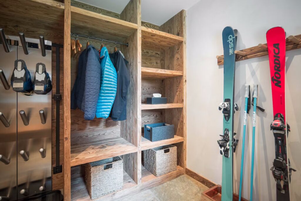 A wooden mudroom with shelves, baskets, hanging jackets, ski boots on racks, and skis leaning against the wall. The space is organized and designed for winter sports gear storage. David Pearson Travel