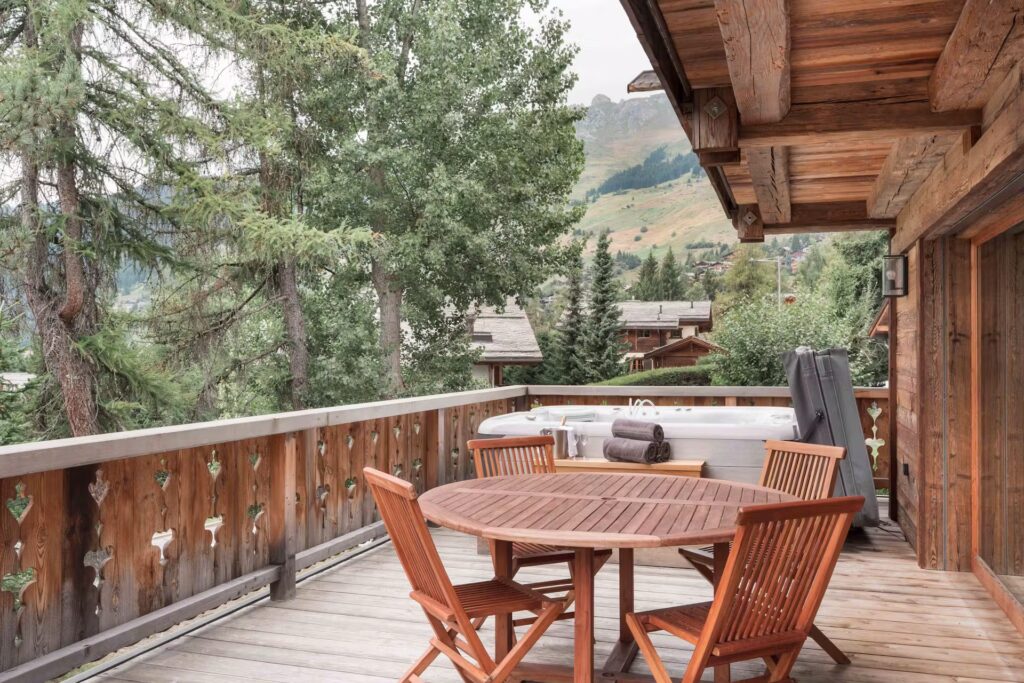 A wooden deck with a round table and chairs overlooks trees, mountains, and chalet-style houses. The deck features a hot tub and rustic railings, creating a cozy, natural atmosphere. David Pearson Travel