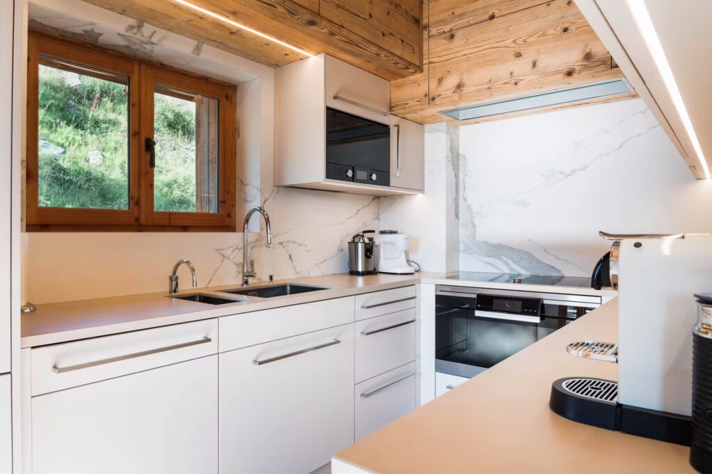 Modern kitchen with white cabinets, marble backsplash, built-in appliances, a sink under a wooden-framed window, and wooden ceiling panels. Natural light enters from the window on the left. David Pearson Travel