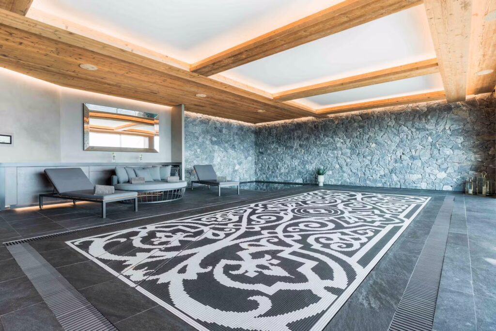 Spacious, modern lounge with stone walls, wooden beam ceiling, skylights, and several gray lounge chairs. The floor features a large, decorative black and white patterned rug. David Pearson Travel