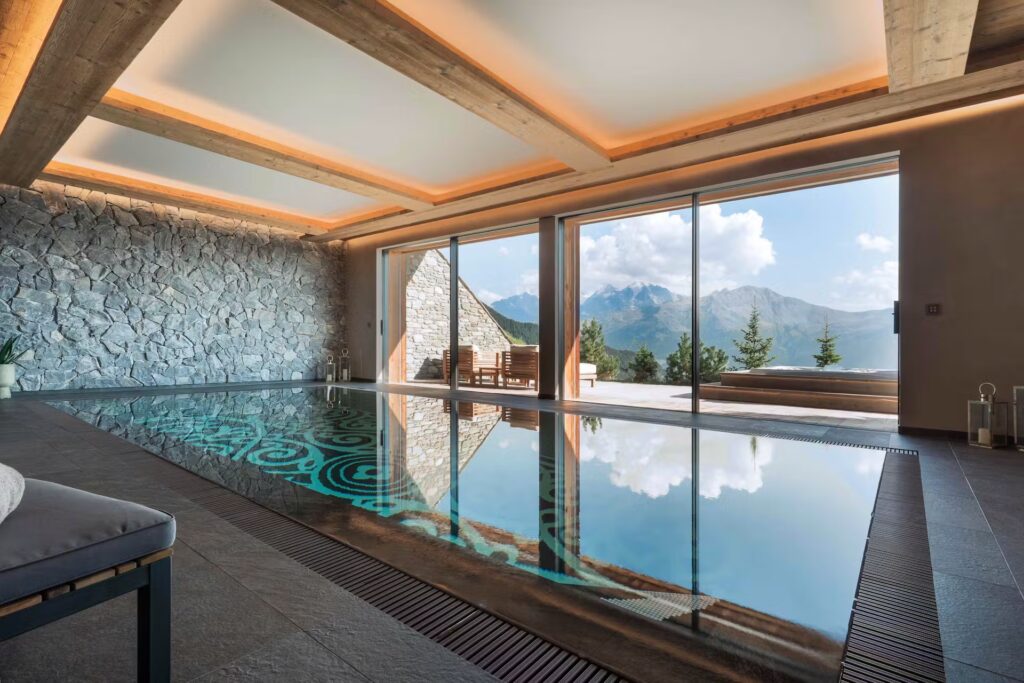 Indoor pool with turquoise patterned tiles, stone wall, and wooden ceiling beams. Large glass doors open to a mountain view with trees and outdoor seating under sunlight. Calm, modern, and inviting atmosphere. David Pearson Travel