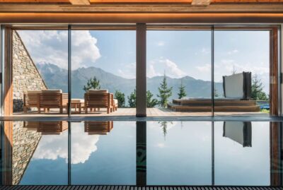 A luxurious indoor pool faces large glass doors opening to a terrace with wooden lounge chairs and a hot tub, overlooking mountains and pine trees under a clear sky. David Pearson Travel