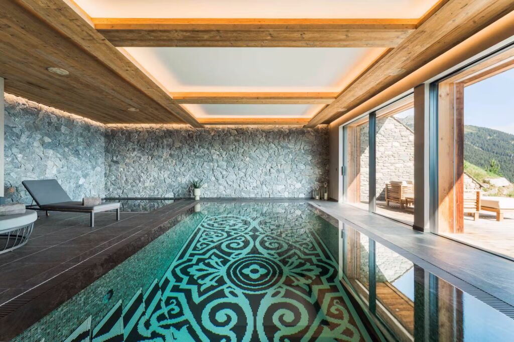 A luxurious indoor pool with ornate tile designs, stone walls, wooden ceiling beams, lounge chairs, and large sliding glass doors opening to a sunny outdoor patio with mountain views. David Pearson Travel