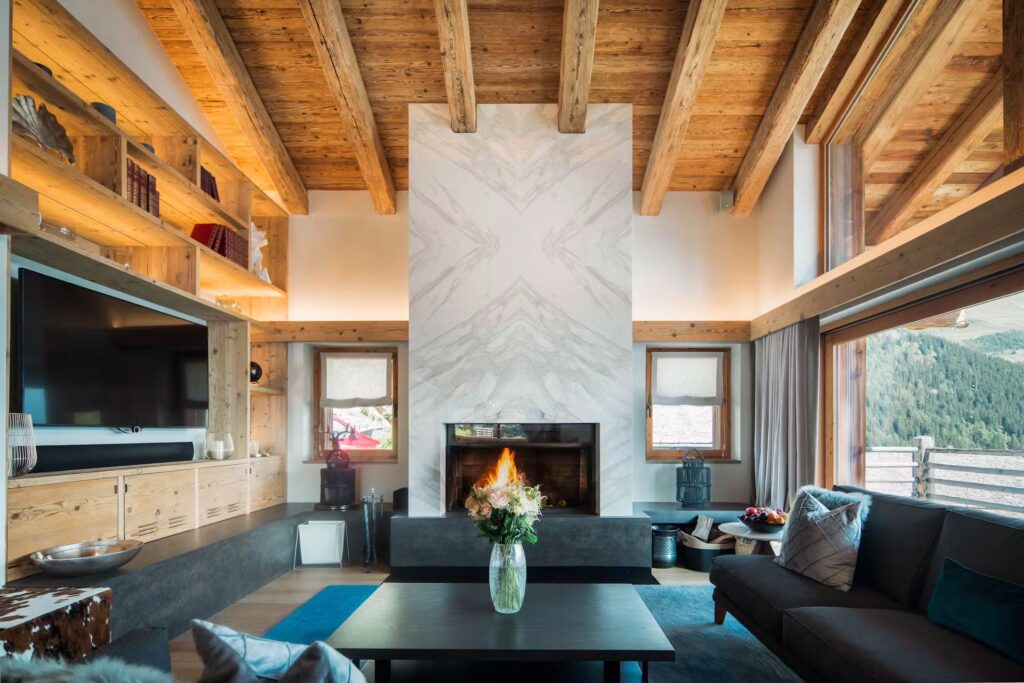 A cozy living room with wood-beamed ceilings, a modern fireplace, large windows with mountain views, built-in shelving, a gray sofa, a coffee table with a vase of flowers, and natural light filling the space. David Pearson Travel