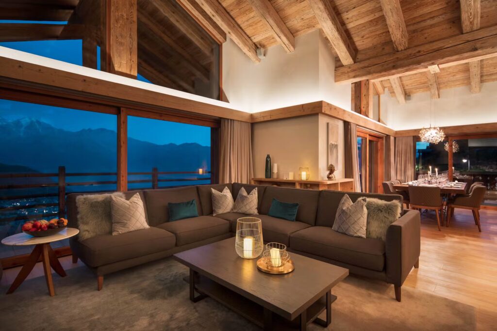 A cozy, modern living room with wooden beams, a large sectional sofa, candlelit coffee table, and floor-to-ceiling windows offering a mountain view at dusk, next to a dining area set for dinner. David Pearson Travel